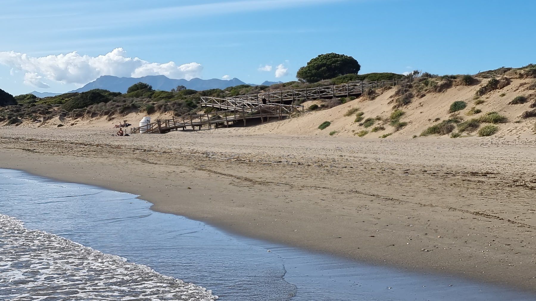 Sandstrand und bewachsene Dünen mit Holzstegen und Bergen im Hintergrund.