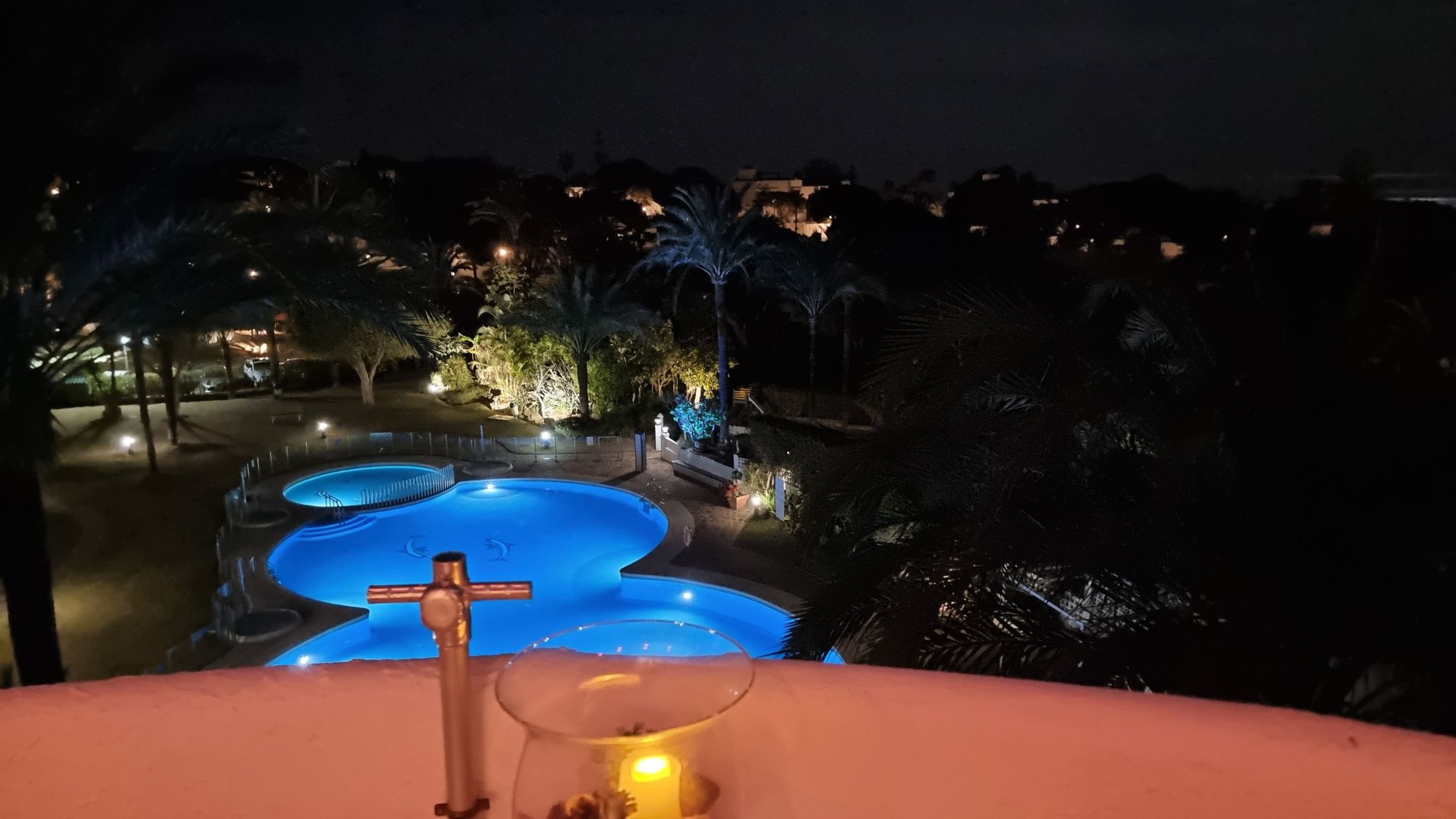 Blau beleuchteter Gemeinschaftspool bei Nacht, davor die Terrassenbrüstung mit einer Kerze